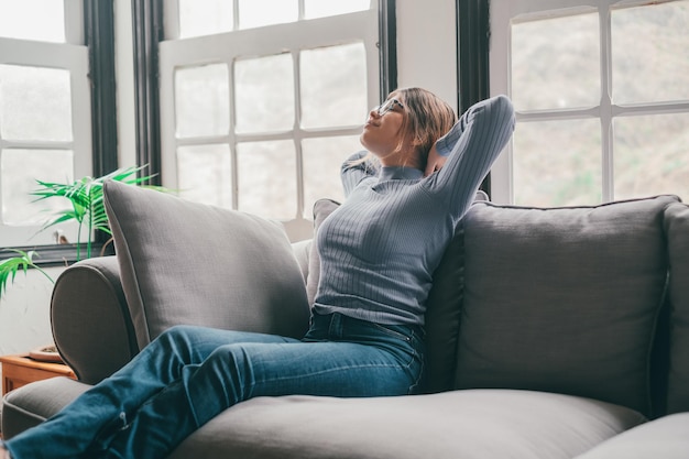 Heureuse jeune femme à lunettes se reposant sur un canapé avec les mains derrière la tête dans le salon de sa maison Dame insouciante et satisfaite de se détendre sur un canapé dans un appartement modernexA