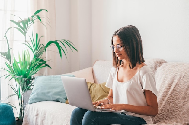 Heureuse jeune femme à lunettes tapant à l'aide d'un ordinateur portable alors qu'elle était assise sur un canapé dans le salon de sa maison. Belle dame passe son temps libre à l'aide d'un ordinateur portable. Indépendant travaillant à domicile