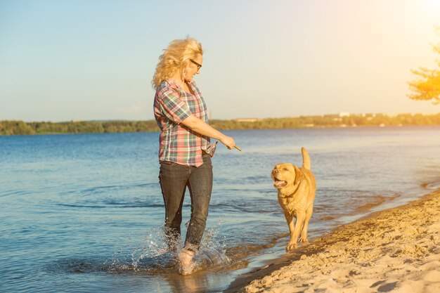 Heureuse jeune femme marchant le long d'une plage avec son golden retriever au coucher de soleil flare