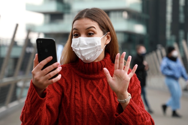 Heureuse jeune femme avec masque chirurgical appel vidéo lors de la marche dans la rue de la ville