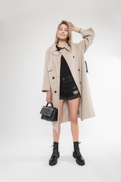 Heureuse jeune femme de mode avec sourire dans des vêtements élégants avec sac à main manteau classique et chaussures noires en cuir pose en studio