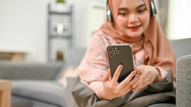 Heureuse jeune femme musulmane asiatique écoutant de la musique avec des écouteurs sur un canapé dans le salon