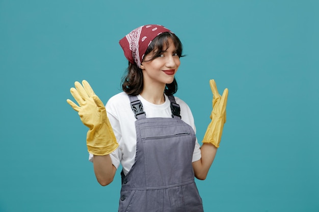 Heureuse jeune femme nettoyante portant un bandana uniforme et des gants en caoutchouc regardant la caméra montrant les mains vides isolées sur fond bleu