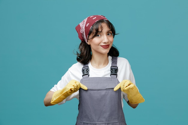 Heureuse jeune femme nettoyante portant un bandana uniforme et des gants en caoutchouc saisissant son uniforme regardant la caméra isolée sur fond bleu