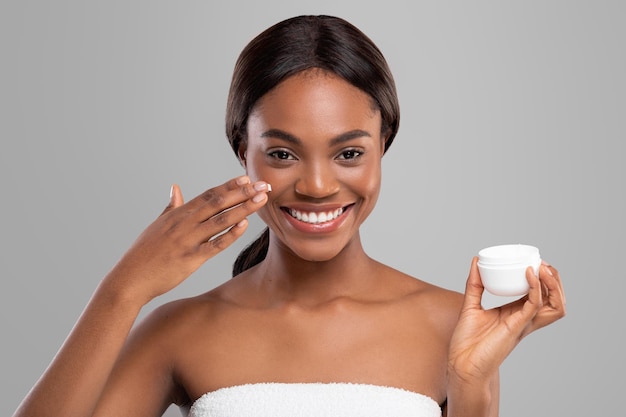 Heureuse jeune femme noire avec une peau parfaite appliquant une crème anti-âge hydratante sur son visage et tenir