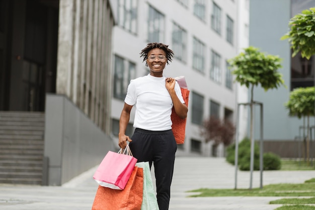 Heureuse jeune femme noire souriante aux cheveux bouclés posant devant la caméra avec des sacs à provisions colorés