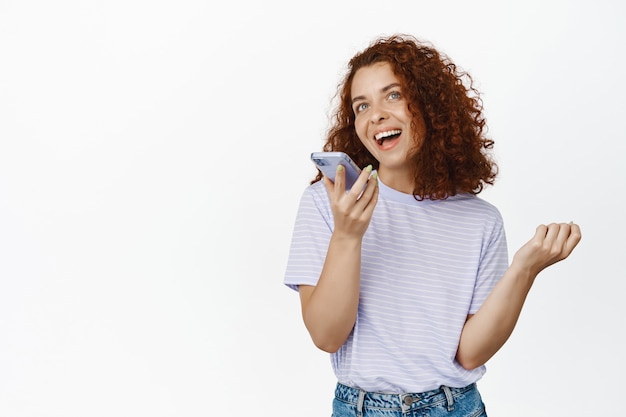 Heureuse jeune femme parlant au téléphone portable, chantant sur un smartphone dynamique, enregistrer la voix sur le haut-parleur, envoyer un message vocal sur blanc.