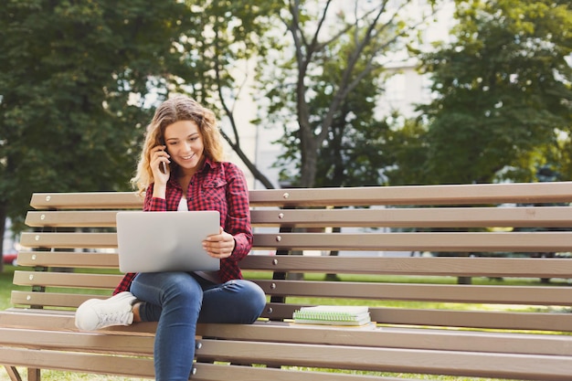 Heureuse jeune femme parlant sur mobile, pigiste travaillant sur ordinateur portable, assise dans une atmosphère agréable à l'extérieur dans le parc. Technologie, communication, éducation et concept de travail à distance, espace de copie