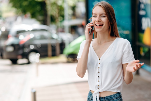 Heureuse jeune femme parlant sur smartphone debout et gesticulant sa main en plein air