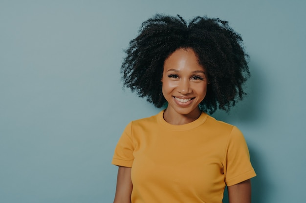 Heureuse jeune femme à la peau foncée avec une coiffure frisée souriant à la caméra avec une expression de visage heureuse, femme africaine en chemise jaune exprimant le bonheur, posant sur fond bleu studio