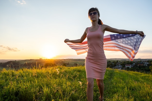 Photo heureuse jeune femme posant avec le drapeau national des états-unis debout à l'extérieur au coucher du soleil.