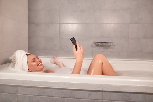 Heureuse jeune femme prenant un bain avec de la mousse et parler par appel vidéo