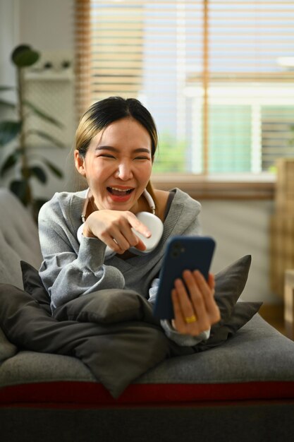 Heureuse jeune femme profitant d'un appel vidéo de chat virtuel en ligne avec des amis via un téléphone portable tout en étant allongé sur un canapé à la maison