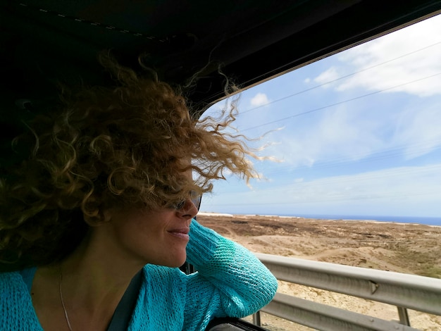 Heureuse jeune femme profitant d'un voyage lors d'un voyage en voiture. Femme à lunettes de soleil à l'extérieur de la fenêtre. Femme aux cheveux ébouriffés admirant la nature depuis la fenêtre de la voiture pendant le voyage sur la route