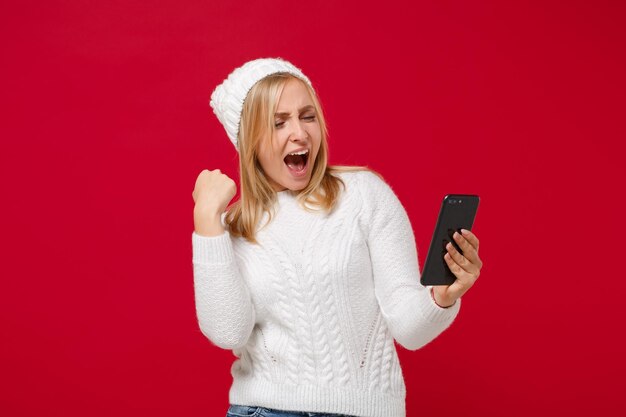 Heureuse jeune femme en pull blanc, chapeau isolé sur fond rouge. Mode de vie sain, concept de saison froide. Maquette de l'espace de copie. À l'aide d'un téléphone portable, en tapant un message sms, en faisant le geste du gagnant.