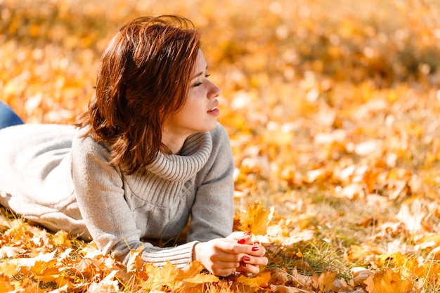 Heureuse jeune femme en pull tricoté se trouve dans le parc sur le feuillage d'automne par une chaude journée ensoleillée ca