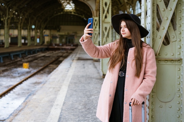 Heureuse jeune femme sur le quai de la gare en manteau rose et chapeau noir attend le train...