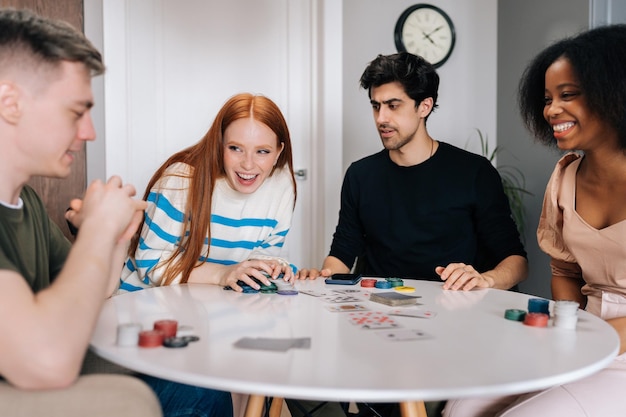 Heureuse jeune femme ravie en riant célébrant la victoire au jeu de poker assis à table dans le salon Groupe d'hommes et de femmes multiethniques s'amusant et profitant de la compétition à la maison