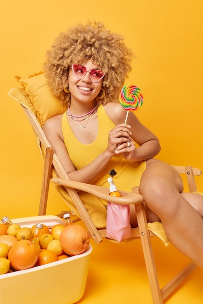 Heureuse jeune femme rêveuse a des cheveux bouclés naturels porte des lunettes de soleil roses et un maillot de bain tient de grandes poses de sucettes multicolores sur une chaise longue près d'un congélateur portable focalisé loin sourit positivement