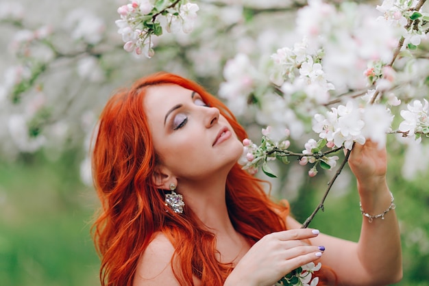 Heureuse jeune femme rousse se dresse dans un verger de pommiers en fleurs, gros plan