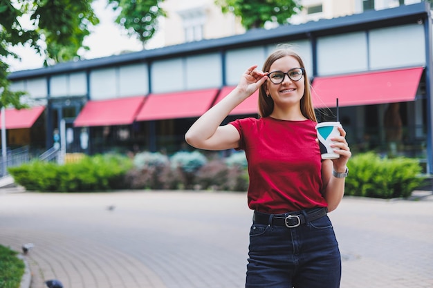 Heureuse jeune femme avec sac à dos souriant et regardant ailleurs tout en marchant dans la rue de la ville et en dégustant du café frais à emporter