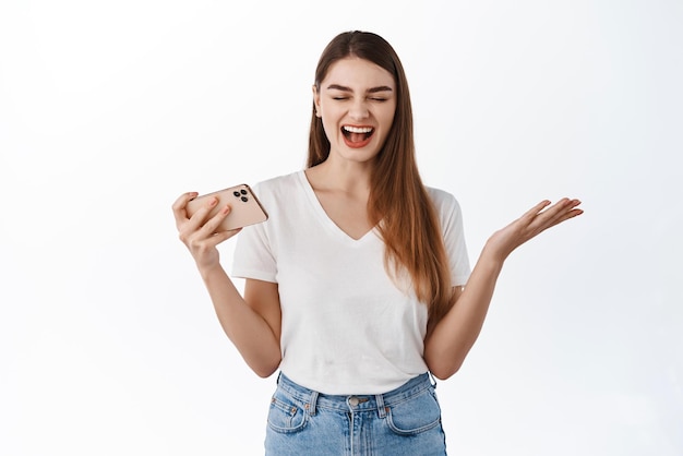 Heureuse jeune femme se réjouissant de gagner sur smartphone en souriant et en criant oui avec joie célébrant la victoire en ligne tenant la victoire horizontale du téléphone dans le jeu vidéo fond blanc