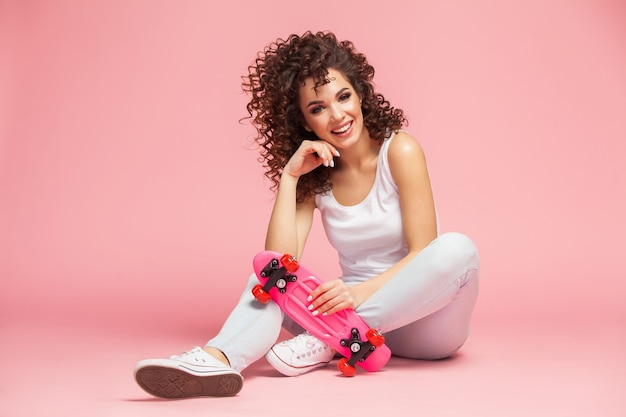 Heureuse jeune femme séduisante assise sur une planche à roulettes sur rose.