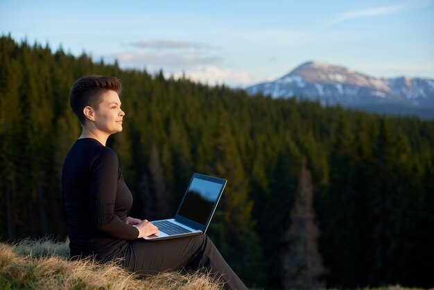 Heureuse jeune femme séduisante travaillant sur son ordinateur portable à l'extérieur, profitant de la vue sur les montagnes