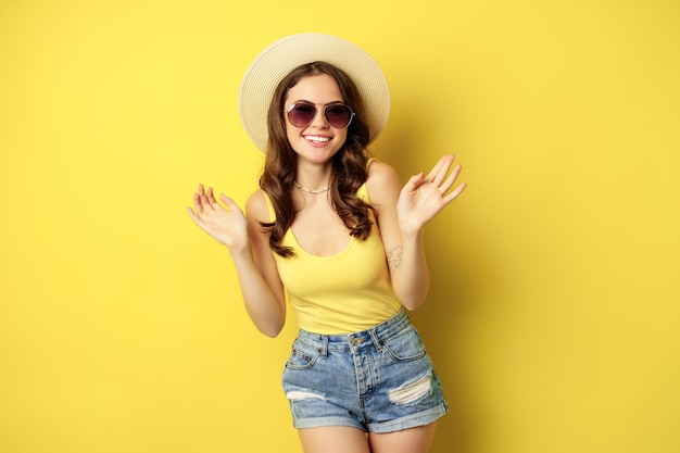 Heureuse jeune femme sexy en vacances, portant un chapeau de paille, des lunettes de soleil, riant et souriant, debout sur fond jaune