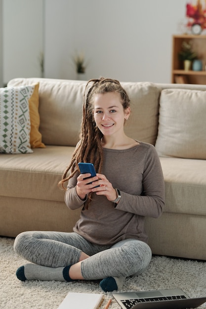 Heureuse jeune femme avec smartphone assis sur le sol par canapé, vous regarde et faire ses devoirs