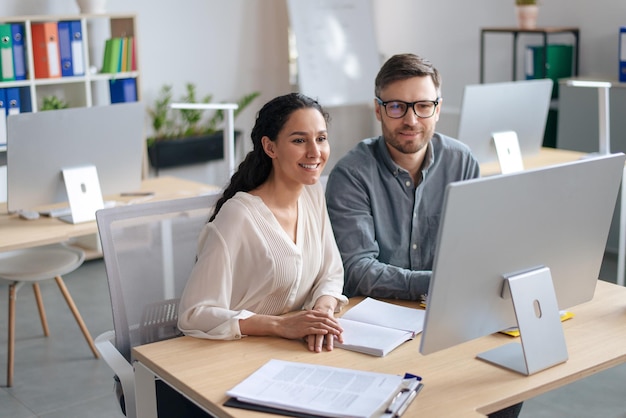 Heureuse jeune femme et son collègue masculin travaillant ensemble à l'aide d'un ordinateur moderne dans un bureau à espace ouvert