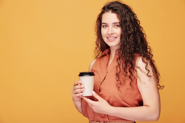 Heureuse jeune femme souriante avec de longs cheveux ondulés noirs tenant un verre de café en position debout avec fond sur la gauche