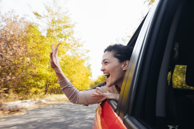 Heureuse jeune femme souriante regardant par la fenêtre de la voiture en déplacement et agitant