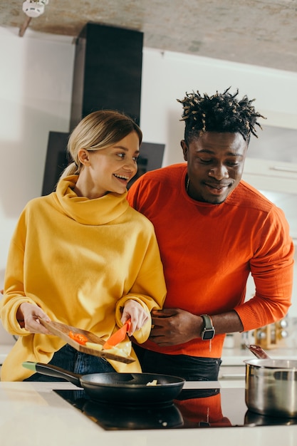 Heureuse jeune femme souriante et regardant son petit ami en se tenant debout avec une planche à découper et des légumes dessus