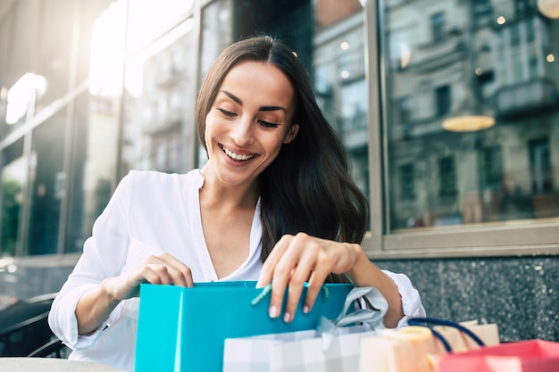 Heureuse jeune femme souriante séduisante regardant dans des sacs à provisions après le centre commercial en plein air