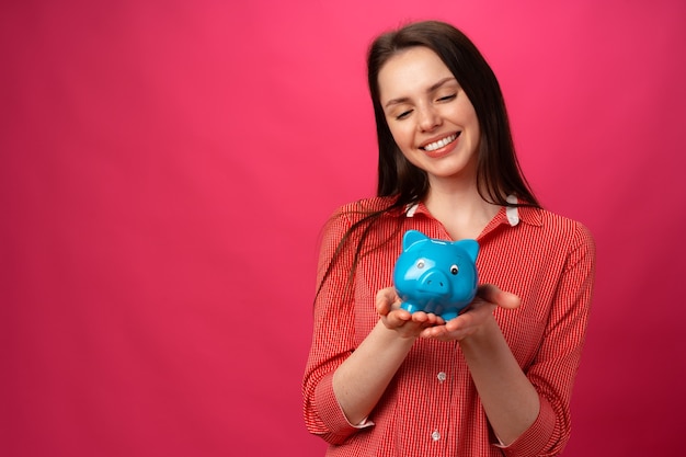 Heureuse jeune femme souriante tenant une tirelire bleue sur fond rose