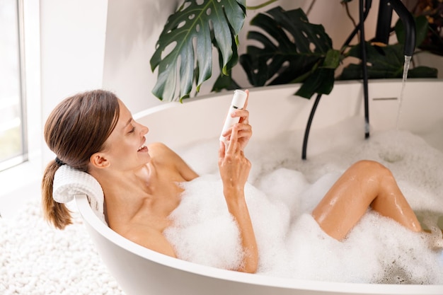 Heureuse jeune femme souriante tout en prenant un bain moussant et en prenant soin de sa peau dans un spa de luxe