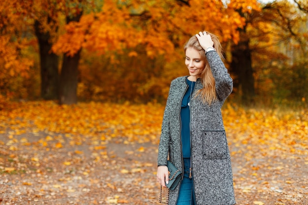 Heureuse jeune femme avec un sourire en vêtements d'automne à la mode posant près des arbres à feuillage jaune