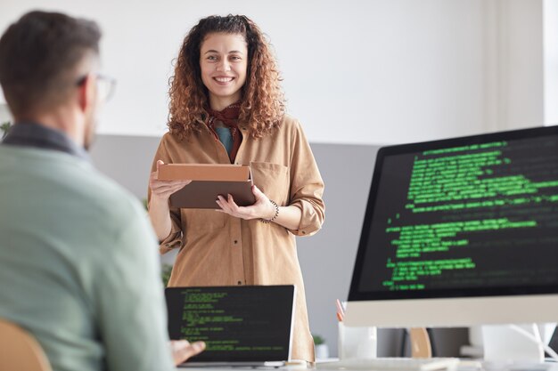 Heureuse jeune femme avec tablette numérique en consultation avec son collègue pendant qu'il travaille sur ordinateur avec logiciel