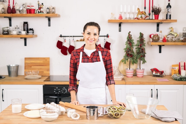 Heureuse jeune femme en tablier debout par table dans la cuisine tout en allant faire des cookies