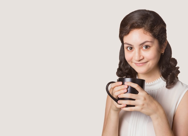 Heureuse jeune femme tenant une tasse de café en céramique sombre, fond de couleur marron clair