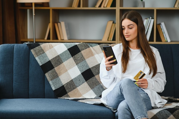 Heureuse jeune femme tenant un téléphone et une carte de crédit assis sur le canapé Achats en ligne