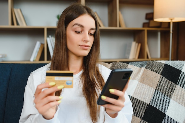 Heureuse jeune femme tenant un téléphone et une carte de crédit assis sur le canapé Achats en ligne