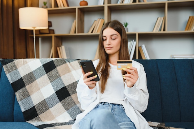 Heureuse jeune femme tenant un téléphone et une carte de crédit assis sur le canapé Achats en ligne