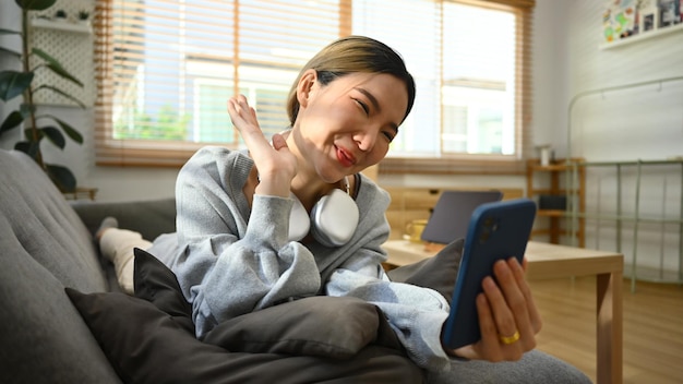 Heureuse jeune femme tenant un téléphone intelligent faisant un appel vidéo en parlant avec un ami en regardant une vidéo en position allongée sur un canapé à la maison
