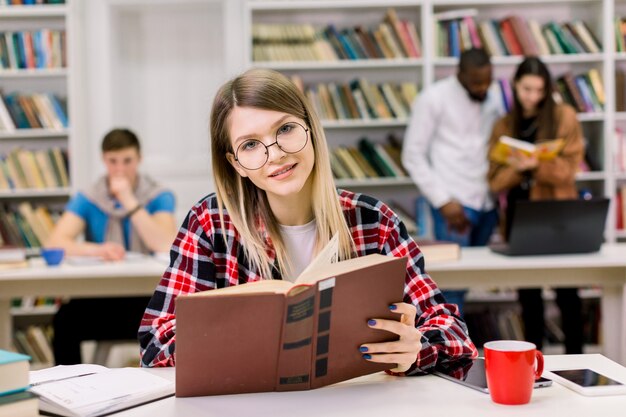 Heureuse jeune femme en tenue décontractée et lunettes assis dans la bibliothèque avec un livre, à la recherche d'informations pour ses études.