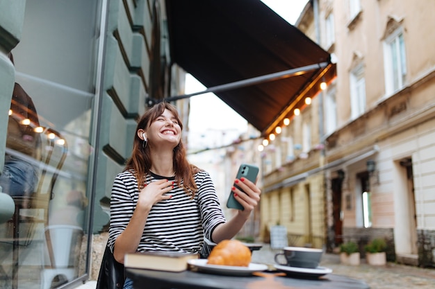 Heureuse jeune femme utilisant un smartphone et des écouteurs ayant un appel vidéo tout en se relaxant sur la terrasse du café