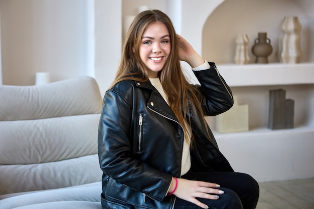 Heureuse jeune femme en veste de cuir est assise sur un canapé à l'intérieur