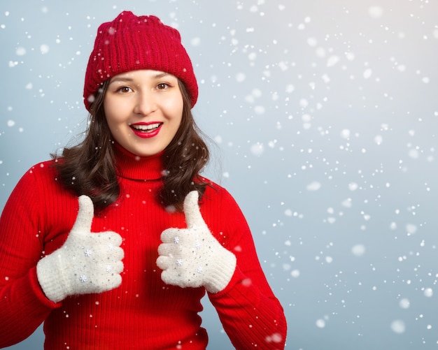Heureuse jeune femme en vêtements tricotés rouges et mitaines montre le geste du pouce vers le haut. contre la surface de la neige qui tombe.