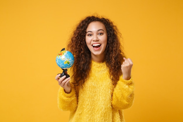 Heureuse jeune fille afro-américaine en pull de fourrure posant isolé sur fond orange jaune, portrait en studio. Concept de style de vie des gens. Maquette de l'espace de copie. Tenant le globe terrestre, faisant le geste du gagnant.